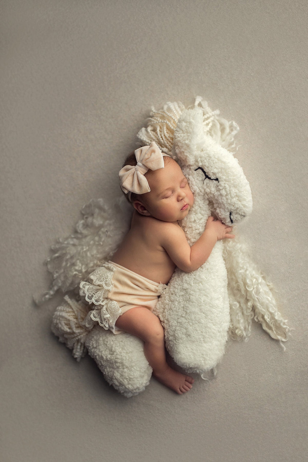 A newborn baby lying next to a white faux fur posing toy designed to resemble a horse, with the baby's arm resting on the toy.