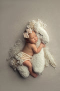 A newborn baby lying next to a white faux fur posing toy designed to resemble a horse, with the baby's arm resting on the toy.