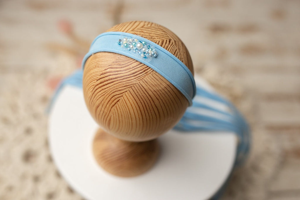 A blue and beige newborn headband displayed on a wooden stand, with a floral pattern on the fabric and a small bead embellishment.