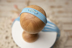A blue and beige newborn headband displayed on a wooden stand, with a floral pattern on the fabric and a small bead embellishment.