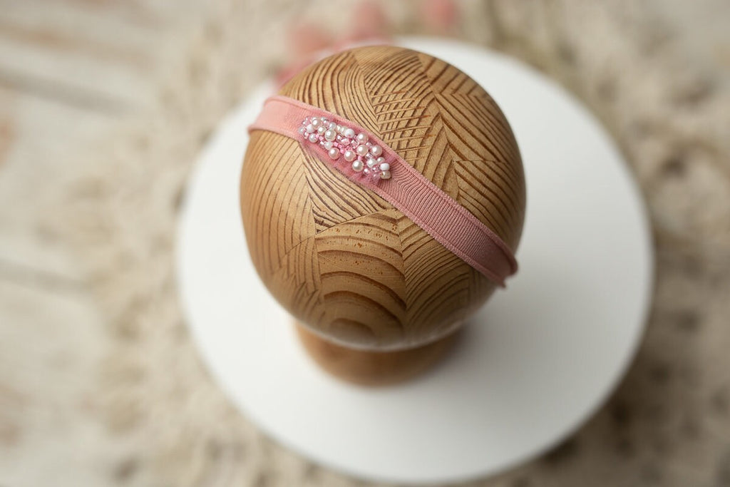 A newborn headband featuring a pink satin bow and bead details, displayed on a wooden mannequin head.